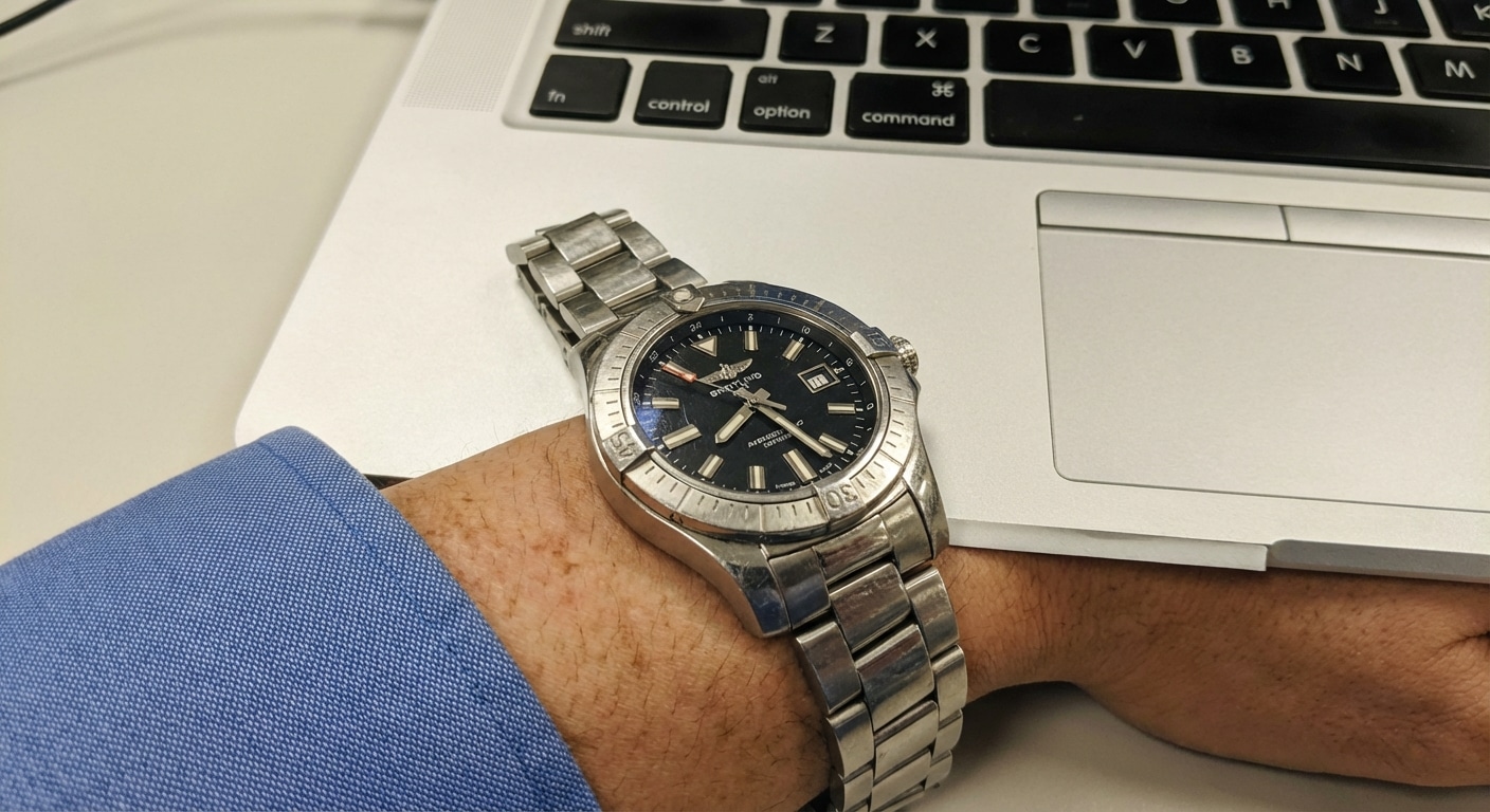 A stainless steel Breitling Avenger replica resting on an aluminum laptop palm rest under harsh office lighting